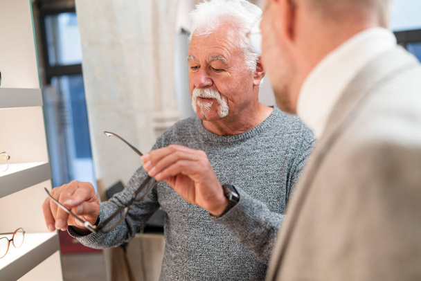 Opticien mène une consultation avec un homme âgé examinant des lunettes dans un magasin d'optique - Photo, image