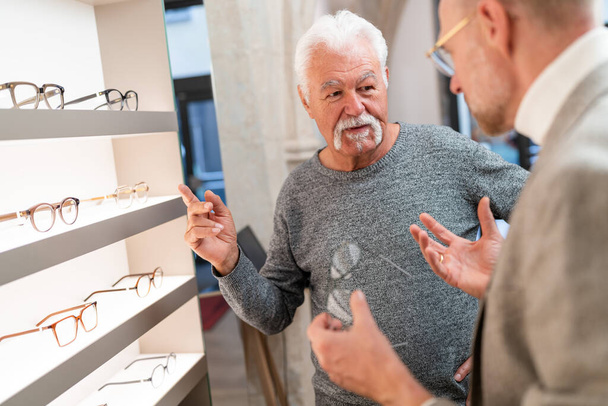 Hombre mayor haciendo gestos durante una conversación con un óptico en una tienda óptica con gafas expuestas en el fondo - Foto, Imagen