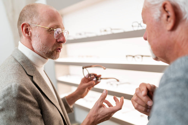 Optician discutindo modelos de óculos com cliente idoso em loja óptica - Foto, Imagem