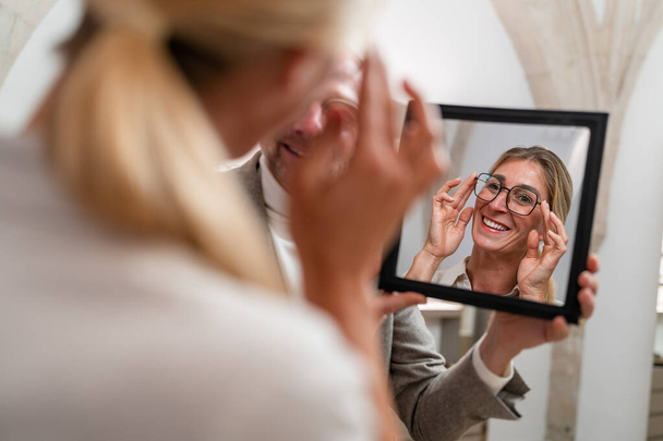 Οπτικός κρατώντας έναν καθρέφτη για να χαμογελά γυναίκα δοκιμάζοντας γυαλιά σε ένα κατάστημα οπτικών. - Φωτογραφία, εικόνα