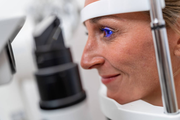 Frau unterzieht sich einer Augenuntersuchung mit Fokus auf ihr beleuchtetes blaues Auge mittels Spaltlampe in der Klinik. Nahaufnahme. Gesundheits- und Medizinkonzept - Foto, Bild