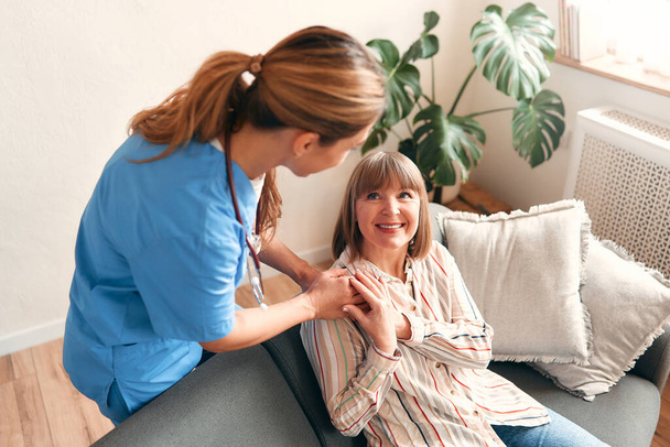 Happy young female nurse provide care medical service help support smiling mature woman at homecare medical visit, lady carer doctor give empathy encourage retired patient sit on sofa at home hospital - Photo, Image