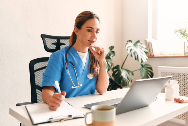 Vrouwelijke arts thuis in medisch uniform met stethoscoop aan tafel met laptop in gesprek op video in de woonkamer. Gezondheidszorg en geneeskunde, online consultatie. - Foto, afbeelding