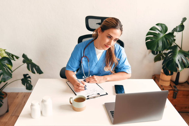 Een arts in een medisch uniform met een stethoscoop aan een tafel met een laptop die aantekeningen maakt en koffie drinkt in het kantoor van een arts in een ziekenhuis. Gezondheidszorg en geneeskunde, online consultatie. - Foto, afbeelding
