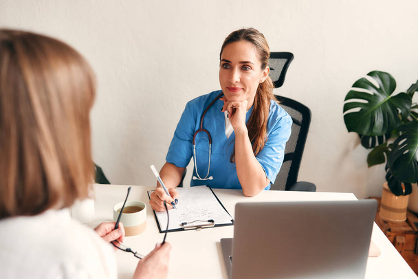 Vrouwelijke arts in medisch uniform met stethoscoop aan tafel met laptop notities maken en praten met de patiënt in het kantoor van de arts in het ziekenhuis. Gezondheidszorg en geneeskunde, online consultatie. - Foto, afbeelding
