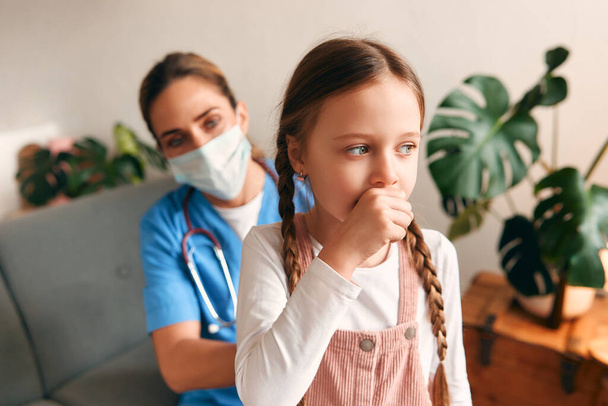 Huisarts thuis in een medisch beschermend masker luisterend naar de longen van een kind terwijl hij op een bank in de woonkamer zit. Patiëntenonderzoek, gezondheidszorg en geneeskunde. - Foto, afbeelding