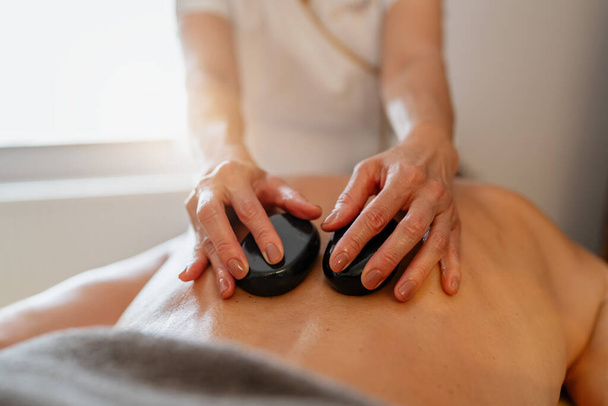 Массажный терапевт, использующий горячие камни на спине клиента во время курортного лечения. Образ отеля Wellness Hotel Concept - Фото, изображение
