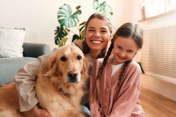 Beautiful mother with cute daughter playing and having fun with dog in cozy living room having a nice time together. - Photo, Image