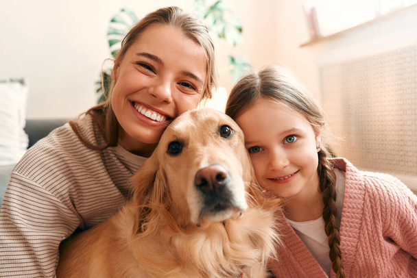 Όμορφη μητέρα με χαριτωμένο κόρη παίζει και να διασκεδάζουν με το σκυλί στο άνετο σαλόνι έχοντας μια ωραία στιγμή μαζί. - Φωτογραφία, εικόνα