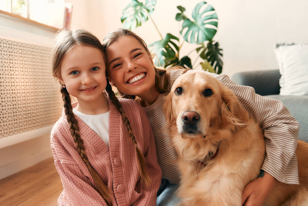 Όμορφη μητέρα με χαριτωμένο κόρη παίζει και να διασκεδάζουν με το σκυλί στο άνετο σαλόνι έχοντας μια ωραία στιγμή μαζί. - Φωτογραφία, εικόνα
