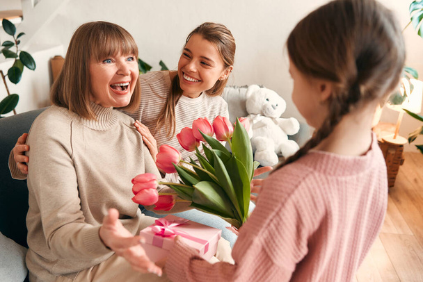 Enkelin und Tochter gratulieren ihrer Großmutter, überreichen ihr Blumen und ein Geschenk, während sie auf dem Sofa im Wohnzimmer sitzen. Konzept von Frauentag und Muttertag. Frauengeneration. - Foto, Bild