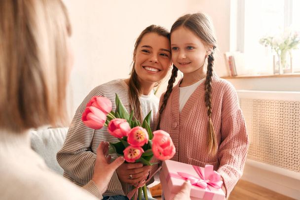 Enkelin und Tochter gratulieren ihrer Großmutter, überreichen ihr Blumen und ein Geschenk, während sie auf dem Sofa im Wohnzimmer sitzen. Konzept von Frauentag und Muttertag. - Foto, Bild