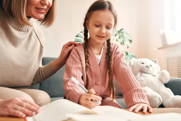 Grandmother and her cute granddaughter are drawing while sitting in the living room on the sofa and having fun. - Photo, Image