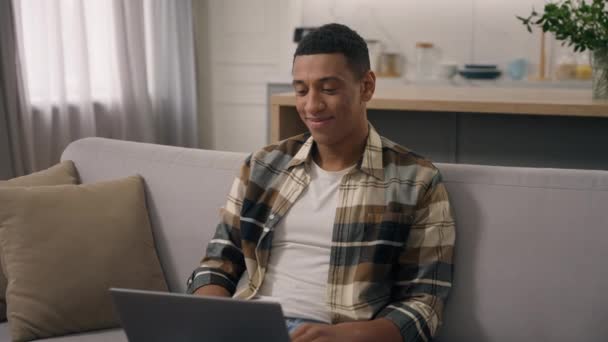 Счастливый улыбчивый афроамериканец этнический фрилансер, работающий дома удаленной онлайн-работой на диване улыбчивый студент, изучающий дистанционно с ноутбуком с помощью компьютерного электронного обучения на диване мужчина с помощью компьютера - Кадры, видео