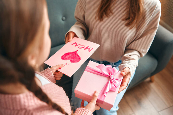 Дитяча дівчинка вітає маму, даруючи їй подарунок і картку, сидячи на дивані у вітальні. День матері і концепція жіночого дня. - Фото, зображення