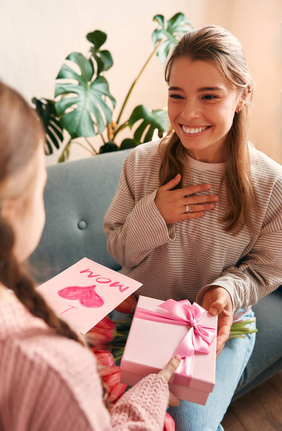 Mädchen gratuliert ihrer Mutter, indem sie ihr ein Geschenk und eine Karte überreicht, während sie auf dem Sofa im Wohnzimmer sitzt. Muttertag und Frauentagskonzept. - Foto, Bild