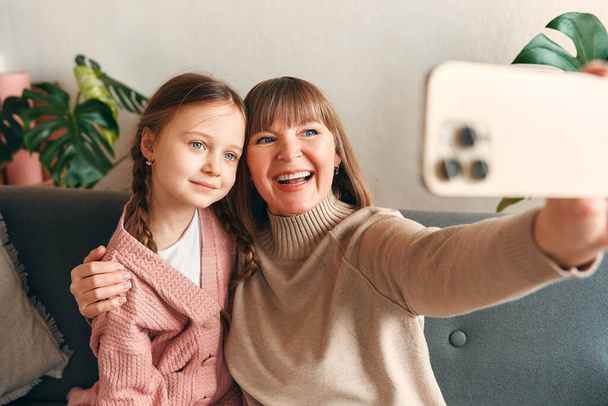 Wnuczka i babcia robią sobie selfie na smartfonie bawiąc się siedząc na kanapie w salonie. Dzień Matki i koncepcja Dnia Kobiet. - Zdjęcie, obraz