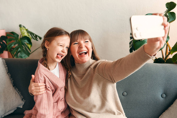 Εγγονή και η γιαγιά λαμβάνοντας μια selfie σε ένα smartphone διασκεδάζοντας ενώ κάθεται στον καναπέ στο σαλόνι. Ημέρα της μητέρας και έννοια ημέρα της γυναίκας. - Φωτογραφία, εικόνα