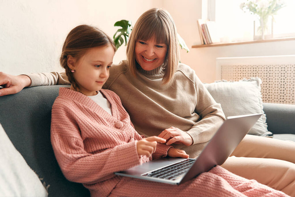 Kleindochter en grootmoeder zitten met een laptop op de bank in de woonkamer en hebben plezier. Moederdag en vrouwendag concept. - Foto, afbeelding