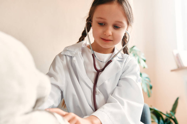 Bambino ragazza in abito medico con uno stetoscopio che tratta un orsacchiotto seduto sul divano in soggiorno. - Foto, immagini