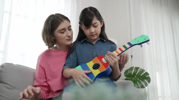 Szczęśliwa dziewczyna gra ukulele podczas gdy kaukaska mama uczy i wyjaśnić o akustycznej muzyki w domu. Słodkie dziecko uczy się o instrumencie. Szczęśliwa, biała matka i dziewczyna spędzają razem czas. Pedagogika. - Materiał filmowy, wideo
