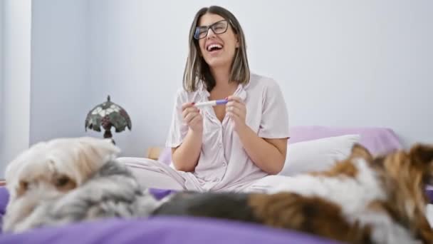 Een vrolijke vrouw in pyjama glimlacht terwijl ze een zwangerschapstest doet in een gezellige slaapkamer geflankeerd door twee honden op haar bed. - Video