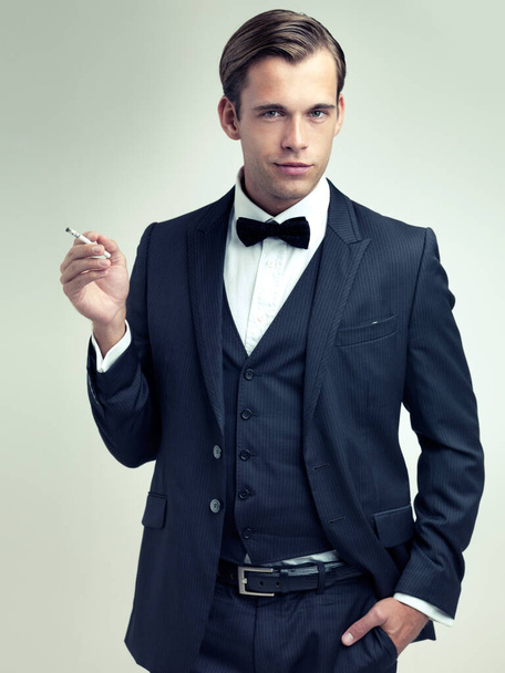 Portrait, elegant or young gentleman smoking a cigarette in studio, confidence or vintage fashion by white background. Face, serious and man in retro suit or tuxedo, bow tie and class at formal event. - Photo, Image