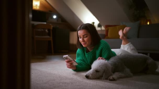 Γκρι σκυλί κοιμάται κοντά στον ιδιοκτήτη του, ενώ η γυναίκα διαβάζει το smartphone της ξαπλωμένη σε μαλακό χαλί στο σπίτι. Έννοια των ανθρώπων και των τεχνολογιών - Πλάνα, βίντεο