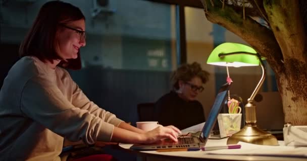 Žena s brýlemi se soustředí na notebook v teple osvětleném večerním kancelářském prostředí, což značí oddanost a práci v pozdních hodinách. - Záběry, video
