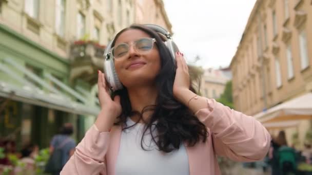 Радостная расслабленная индийская арабская этническая молодежь Z студентка женщина туристка наслаждается прослушиванием музыкального плейлиста приложение танцевальная песня беспроводные наушники звук ритм жизни на улице города на открытом воздухе - Кадры, видео