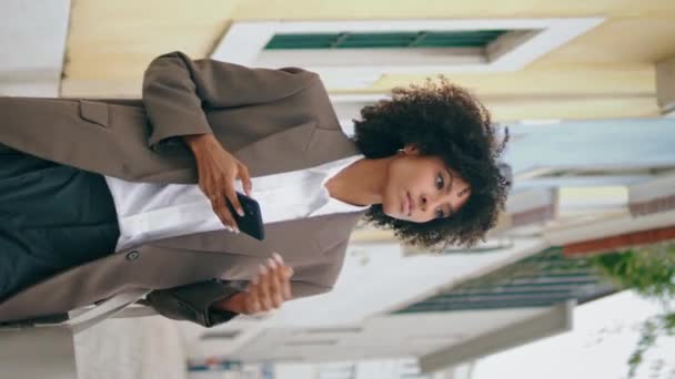 Frau mit Smartphone zu Fuß in stilvollem Anzug Stadt Straße senkrecht ausgerichtet. Schöne afrikanisch-amerikanische Mädchen, die auf dem Telefonbildschirm Tippnachricht. Glückliche lockige Geschäftsfrau, die mit dem Handy zur Arbeit geht. - Filmmaterial, Video