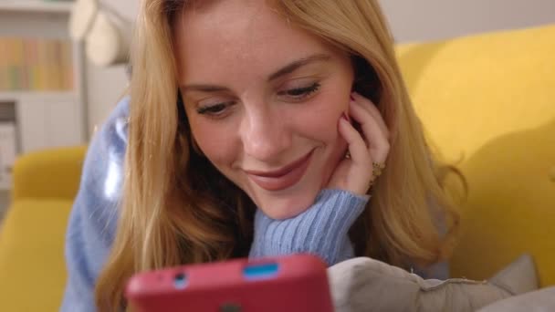 Νεαρή ελκυστική ξανθιά γυναίκα που χρησιμοποιεί το κινητό τηλέφωνο συσκευή, ενώ ξαπλώνουν σε κίτρινο καναπέ στο σπίτι. Έννοια των μέσων κοινωνικής δικτύωσης και τεχνολογίας. - Πλάνα, βίντεο