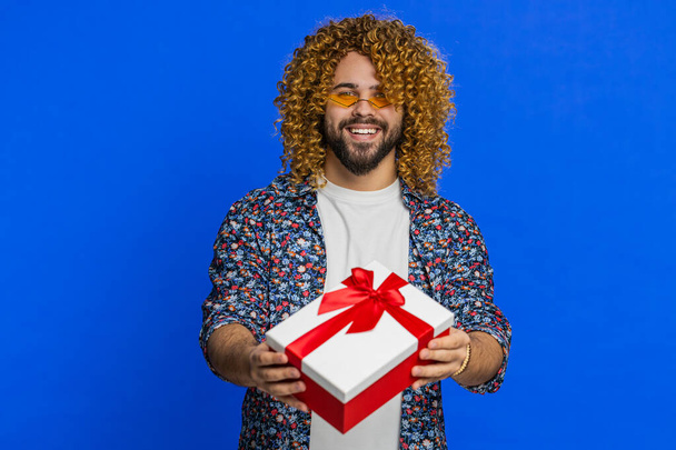 Positive smiling Caucasian man presenting birthday gift box stretches out hands offer wrapped present career bonus celebrating party promotion discount sale. Stylish guy isolated on blue background - 写真・画像