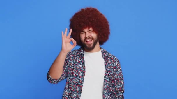 Już idę. Szczęśliwy człowiek z afro peruką fryzurę patrząc z aprobatą na aparat pokazujący ok gest, jak znak pozytywnej opinii, zatwierdzić coś dobrego, świętować zwycięstwo. Facet izolowany na niebieskim tle - Materiał filmowy, wideo