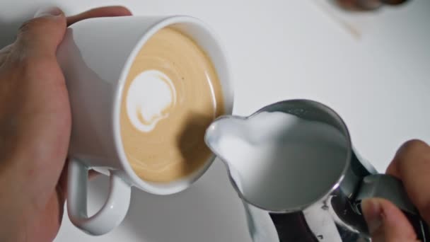 Männerhände machen Latte Art im Coffeeshop in Großaufnahme. Unbekannter Barista schüttet geschlagene Milch in aromatischen Kaffee und schafft so den perfekten Cappuccino-Senkrechtschuss. Café-Mitarbeiterin bastelt Herzmuster an leckerem Getränk - Filmmaterial, Video