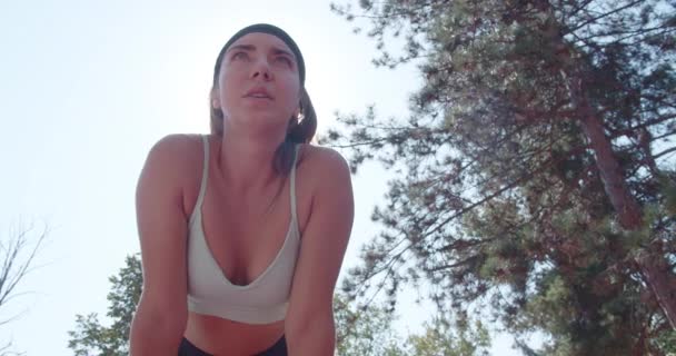 Actieve vrouw in sportkleding rust na een run in een natuurlijke omgeving, vangt haar adem met bomen en lucht boven haar hoofd. - Video