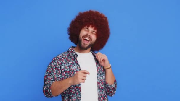 Trendy cheerful positive man with Afro hairstyle wig having fun dancing and moving to rhythm, dabbing raising hands making dub dance winner celebrating victory gesture. Guy isolated on blue background - Footage, Video