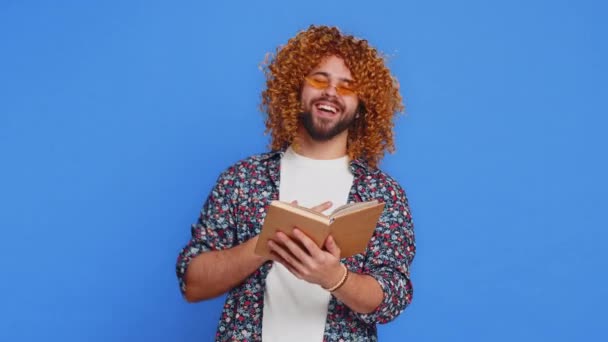 Счастливый расслабленный улыбчивый мужчина с кудрявым париком, читающий смешную интересную сказку, веселый хобби, знания мудрости, образования, обучения, учебы, воя. Парень изолирован на синем фоне - Кадры, видео