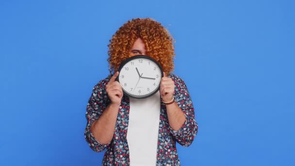 Serious strict man with curly hairstyle wig holding office clock hiding behind, checking time on watch, running late to work, being in delay, deadline, obscuring face. Guy isolated on blue background - Footage, Video