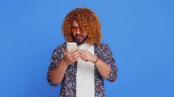 Upset sad man with curly hairstyle wig use smartphone typing browsing, play game, loses, surprised by sudden lottery results bad fortune loss fail unlucky news. Stylish guy isolated on blue background - Footage, Video