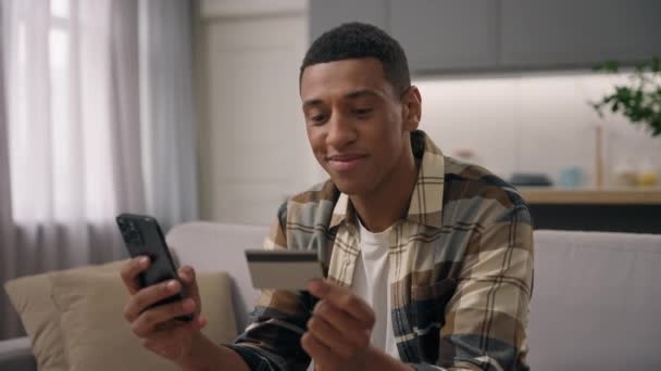 Χαμογελώντας Αφροαμερικανός αγοραστής μακρινό πελάτη χρήστη πληρώσει online πληρωμή internet τραπεζική απομακρυσμένη αγορά με κινητό τηλέφωνο ψώνια αγορά παράδοση αγαθών παραγγείλετε ανέμελη αρσενικό έθνικ τύπος στο σπίτι buy - Πλάνα, βίντεο