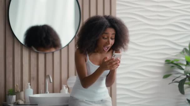 Emocionado logro femenino en casa con teléfono inteligente increíble resultado de noticias ofrecen feliz mujer afroamericana teléfono móvil victoria éxito gritos regocijo celebrar ganar en línea en el baño - Imágenes, Vídeo