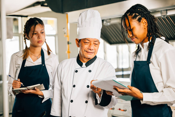 Asian senior chef educates multiracial students in a restaurant kitchen. Emphasizing teamwork learning and note-taking. Professional training in progress. Food Edocation - Fotoğraf, Görsel