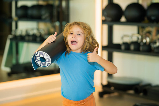 Excited kid with thumbs up holding yoga mat in gym. Yoga child concept. Young strong sporty kid. Workout sport concept - Photo, image
