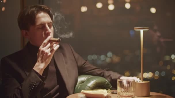 Молодий чоловік в чорному одязі палить сигару в темному ресторані. Медіа. Надійний чоловік дим видихає  - Кадри, відео