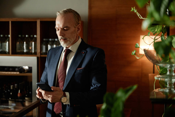 カフェに立っている間にスマートフォンにタイピングメッセージを入力するエレガントなスーツの成熟したビジネスマン - 写真・画像