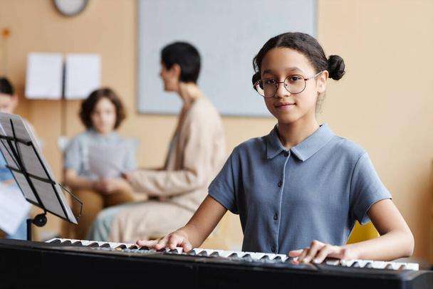 Portrait of schoolgirl looking at camera while learning to play piano in music class - Photo, Image