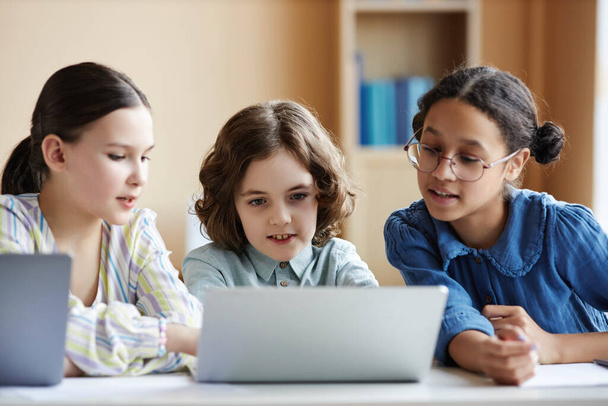 Dzieci w wieku szkolnym korzystające z laptopa do wspólnego oglądania filmów podczas siedzenia przy stole w klasie - Zdjęcie, obraz