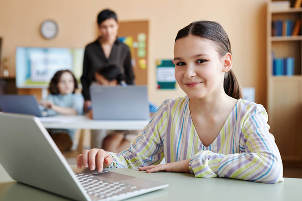 Retrato de colegiala con portátil mirando a la cámara mientras está sentada en la lección con el profesor en segundo plano - Foto, imagen