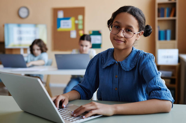 Portrét afroamerické školačky v brýlích, usmívající se na fotoaparát, sedící u stolu a používající notebook během hodiny - Fotografie, Obrázek